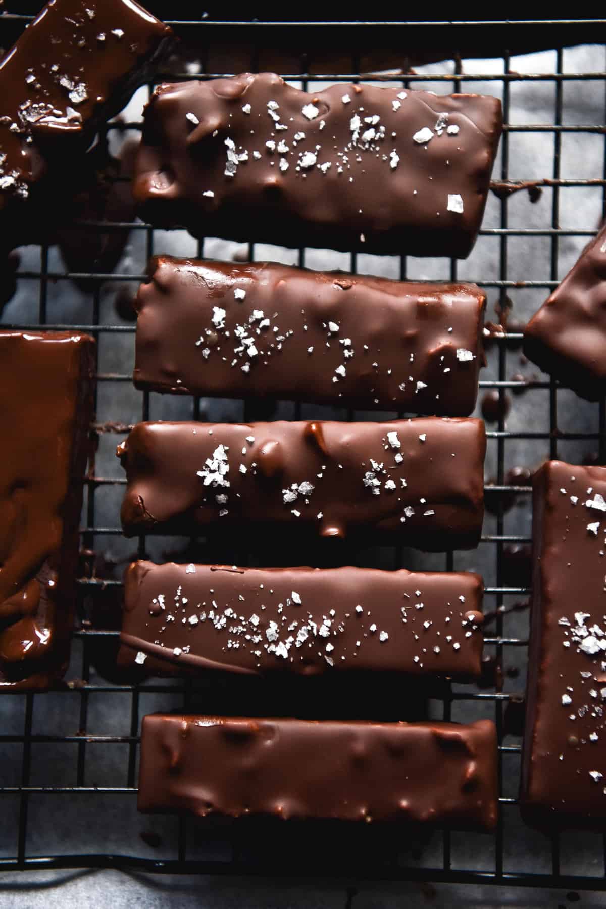 An aerial image of chewy peanut butter bars drying on a baking rack. Their shiny chocolate shells are topped with sea salt flakes and encase toasted peanuts and a peanut centre. They sit atop a cooling rack over a piece of parchment paper.