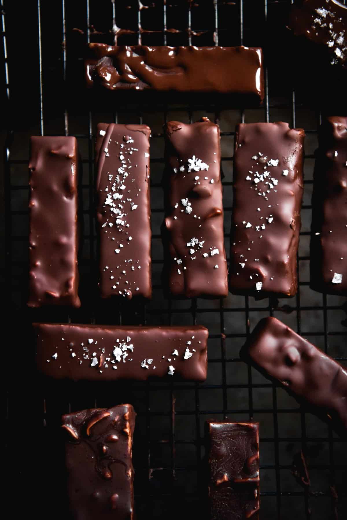 A moody, aerial image of chewy peanut butter bars with sea salt and a dark chocolate coating. The bars sit atop a cooling rack as the chocolate drips off them onto the black background below.