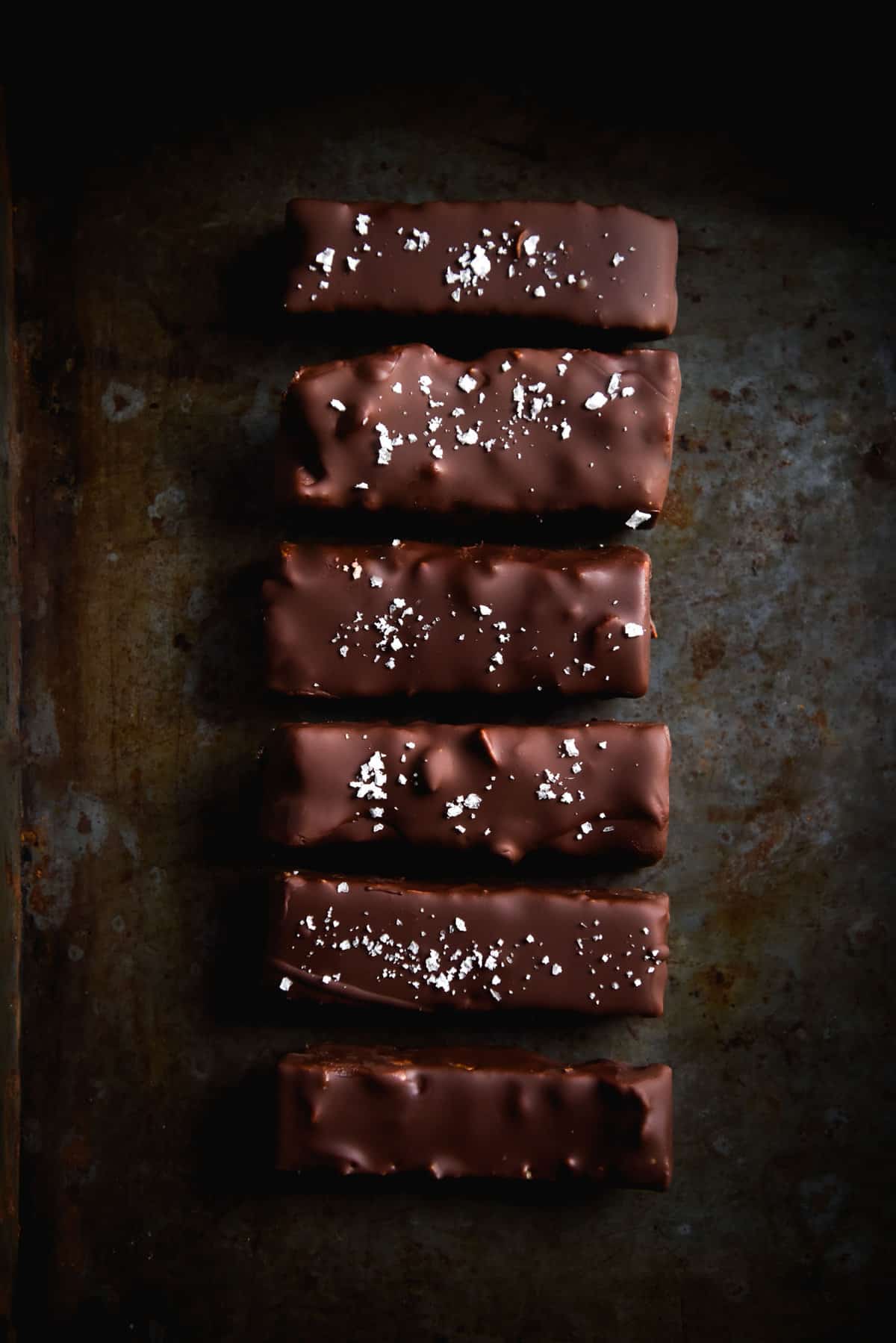 A line of chewy peanut butter bars coated in chocolate and finished with sea salt flakes atop a dark steel backdrop.