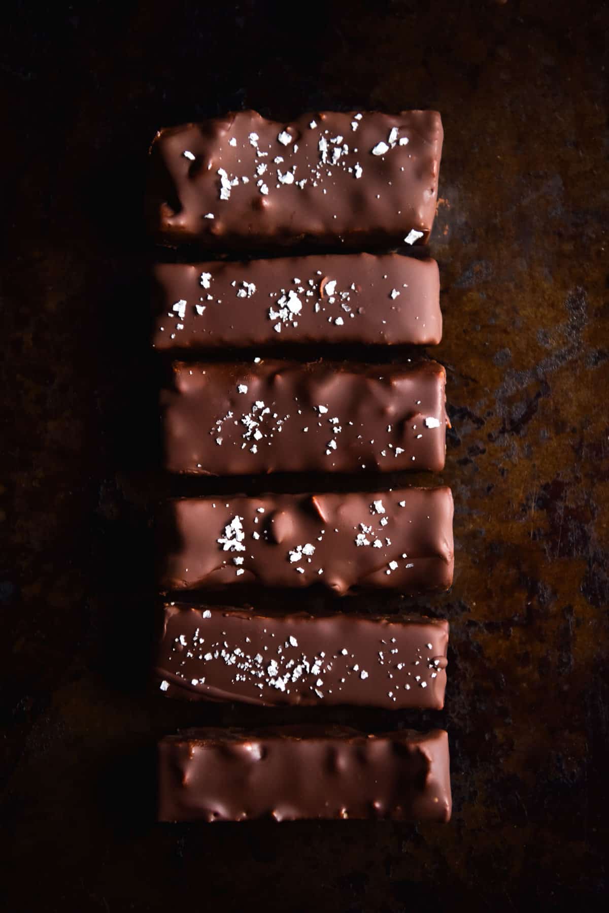 An aerial image of a line of vegan peanut butter chocolate covered bars on a rust coloured backdrop. The bars are chocolate brown in colour and finished with sea salt flakes.