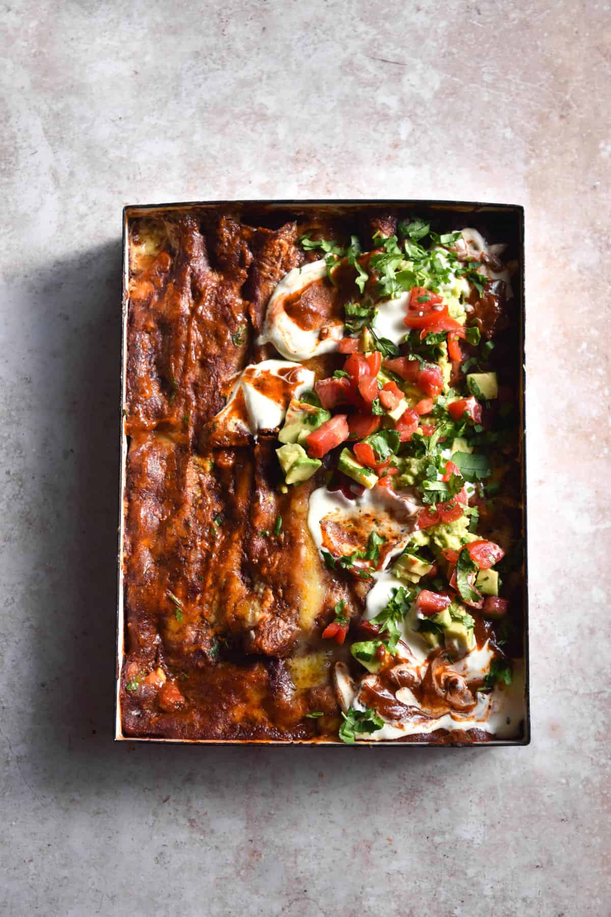 An aerial image of a white baking dish filled with vegetarian enchilada bake. Half of the bake is covered in an avocado and tomato salsa. The baking dish sits atop a light pink backdrop