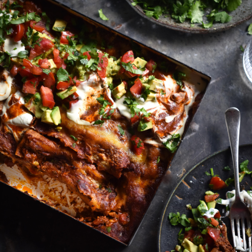 An aerial view of a tray of vegetarian, FODMAP friendly enchiladas topped with sour cream and a tomato, avocado and coriander salsa. The tray sits at an angle on a dark backdrop and is surrounded by dark ceramic plates topped with enchiladas or seasonings and lime wedges.