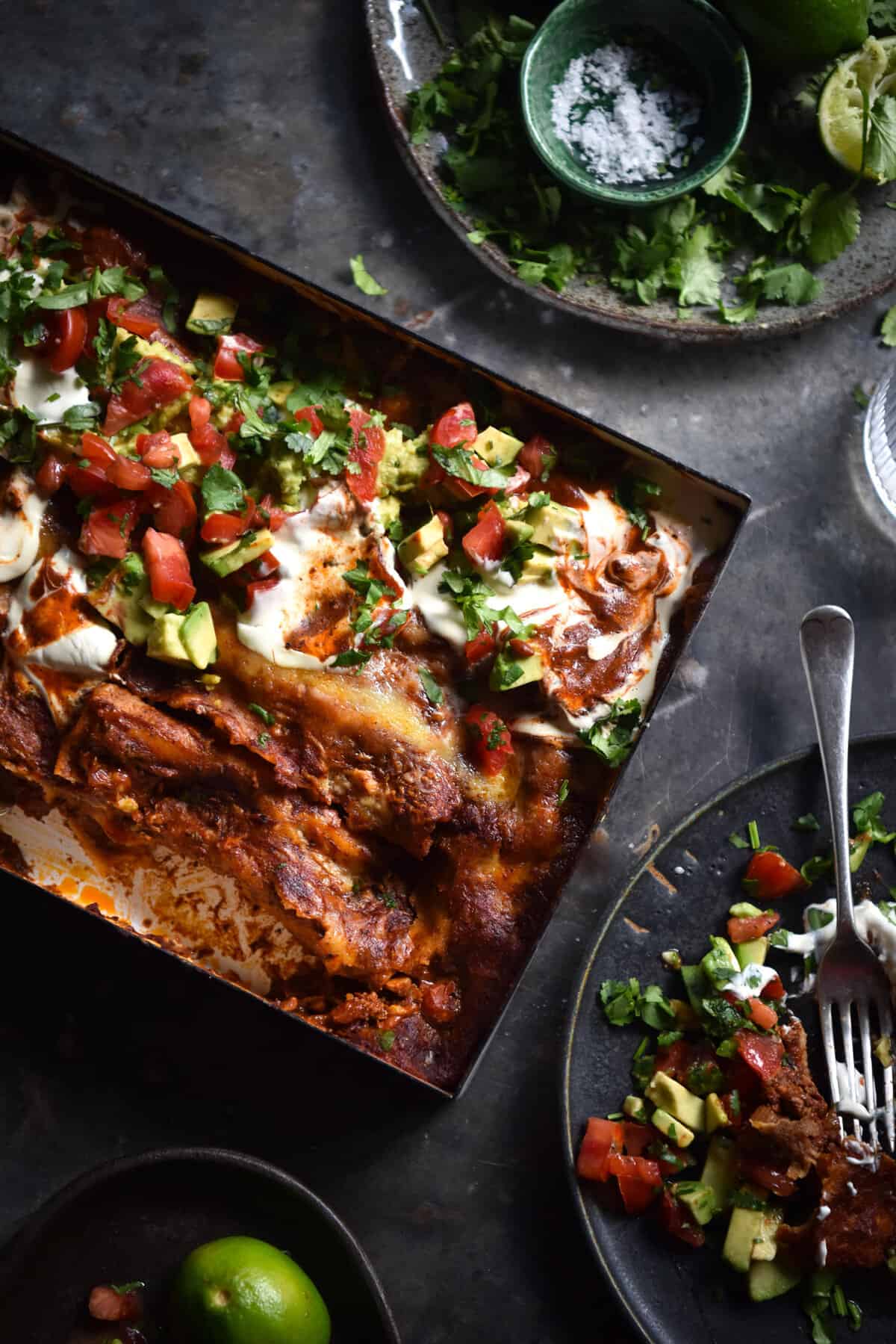 An aerial view of a tray of vegetarian, FODMAP friendly enchiladas topped with sour cream and a tomato, avocado and coriander salsa. The tray sits at an angle on a dark backdrop and is surrounded by dark ceramic plates topped with enchiladas or seasonings and lime wedges.
