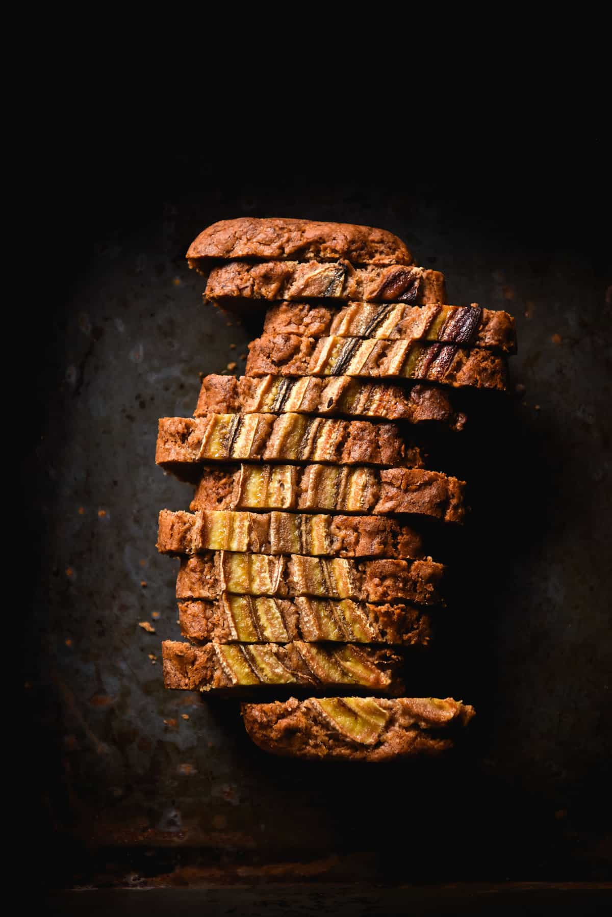 A dark and moody aerial image of a loaf of gluten free vegan banana bread that has been sliced and sits atop a dark steel backdrop