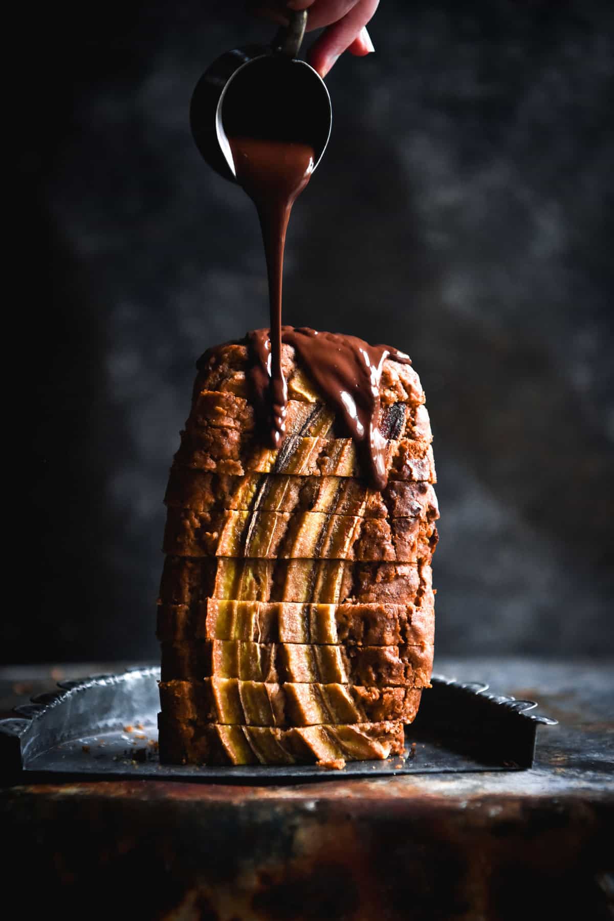 A side on view of a sliced loaf of low FODMAP banana bread standing upright against a dark blue backdrop. A hand extends out from the top right of the image to drizzle vegan chocolate sauce down the loaf.