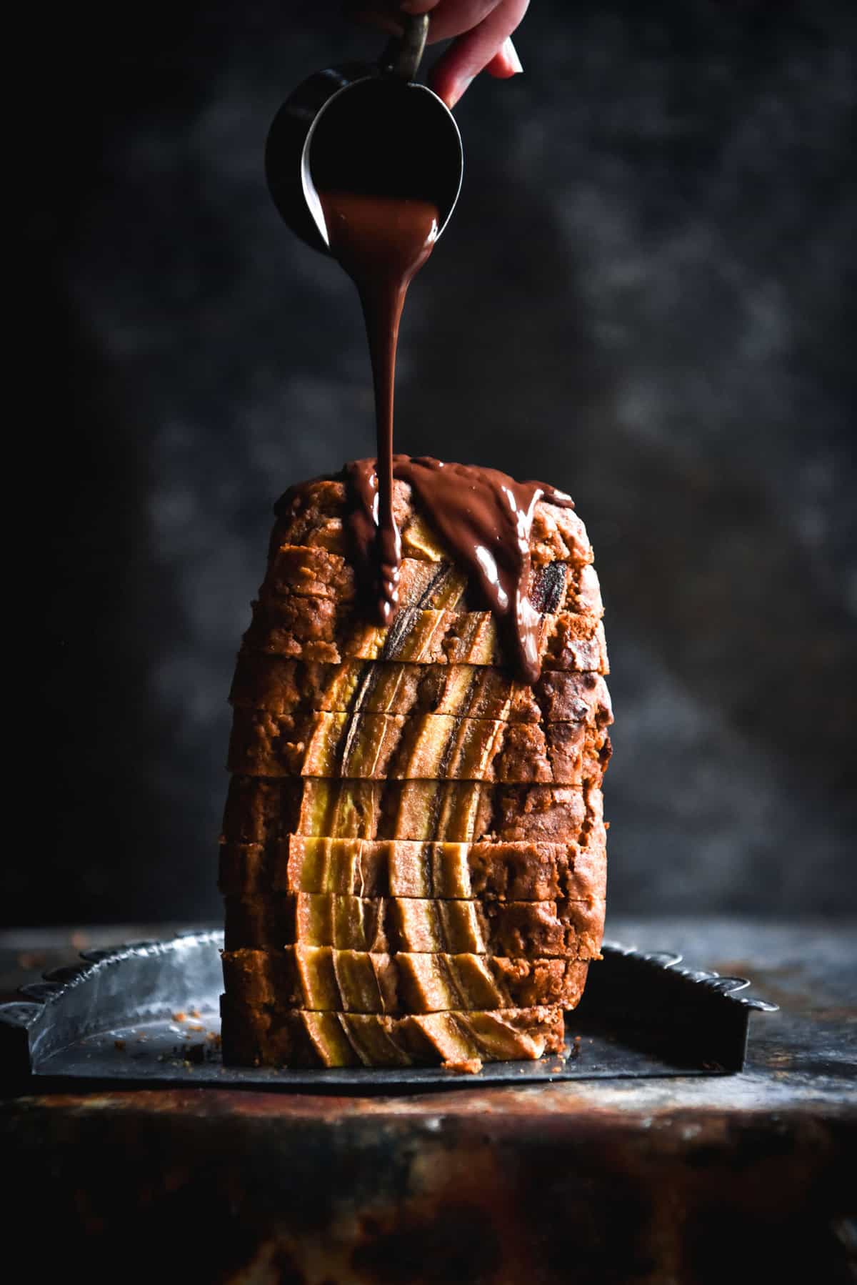 A side on view of a sliced loaf of banana bread standing upright against a dark blue backdrop. A hand extends out from the top right of the image to drizzle vegan chocolate sauce down the loaf.