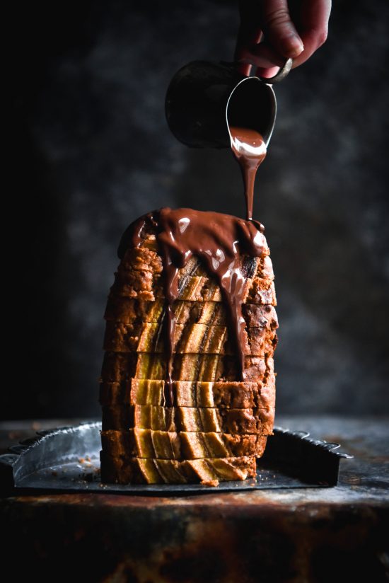 A side on view of a sliced loaf of banana bread standing upright against a dark blue backdrop. A hand extends out from the top right of the image to drizzle vegan chocolate sauce down the loaf.