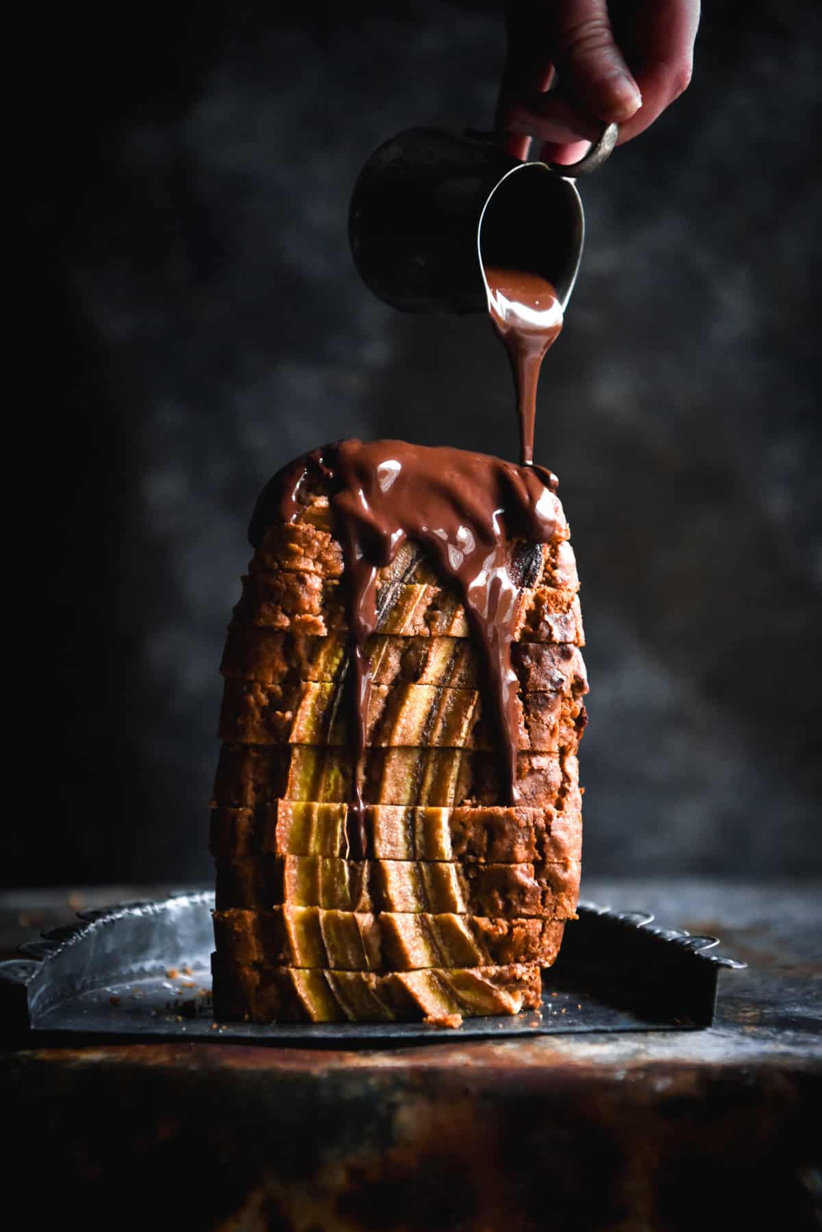 A side on view of a sliced loaf of banana bread standing upright against a dark blue backdrop. A hand extends out from the top right of the image to drizzle vegan chocolate sauce down the loaf.