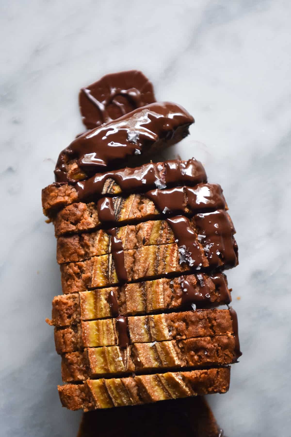 An aerial image of a loaf of vegan, gluten free banana bread that has been sliced and drizzled with chocolate. The loaf sits on a white marble table. 