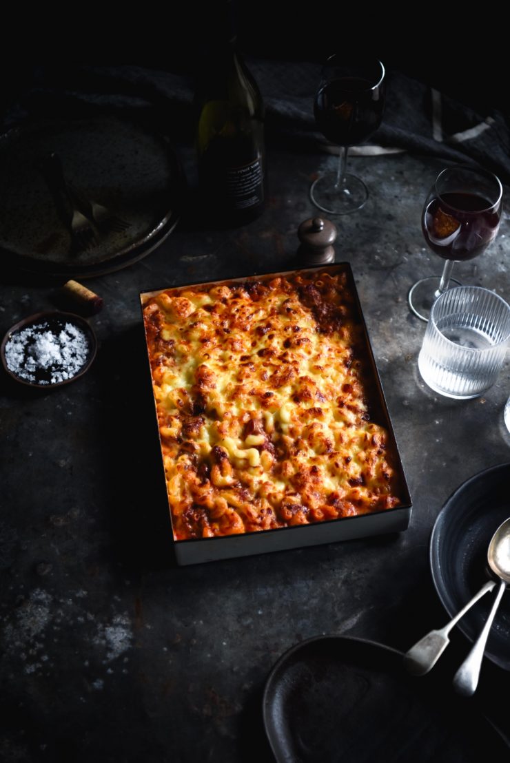 A dark and moody image of a vegetarian hamburger helper bake on a dark grey backdrop. The hamburger helper sits in white rectangular baking dish and is golden brown on top. It is surrounded by glasses of water, plates and cutlery.