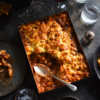 Gluten free, vegetarian bolognese, mac and cheese bake on a dark moody backdrop surrounded by water glasses. A scoop of bake has been removed, revealing the rich vegetarian bolognese underneath the mac and cheese