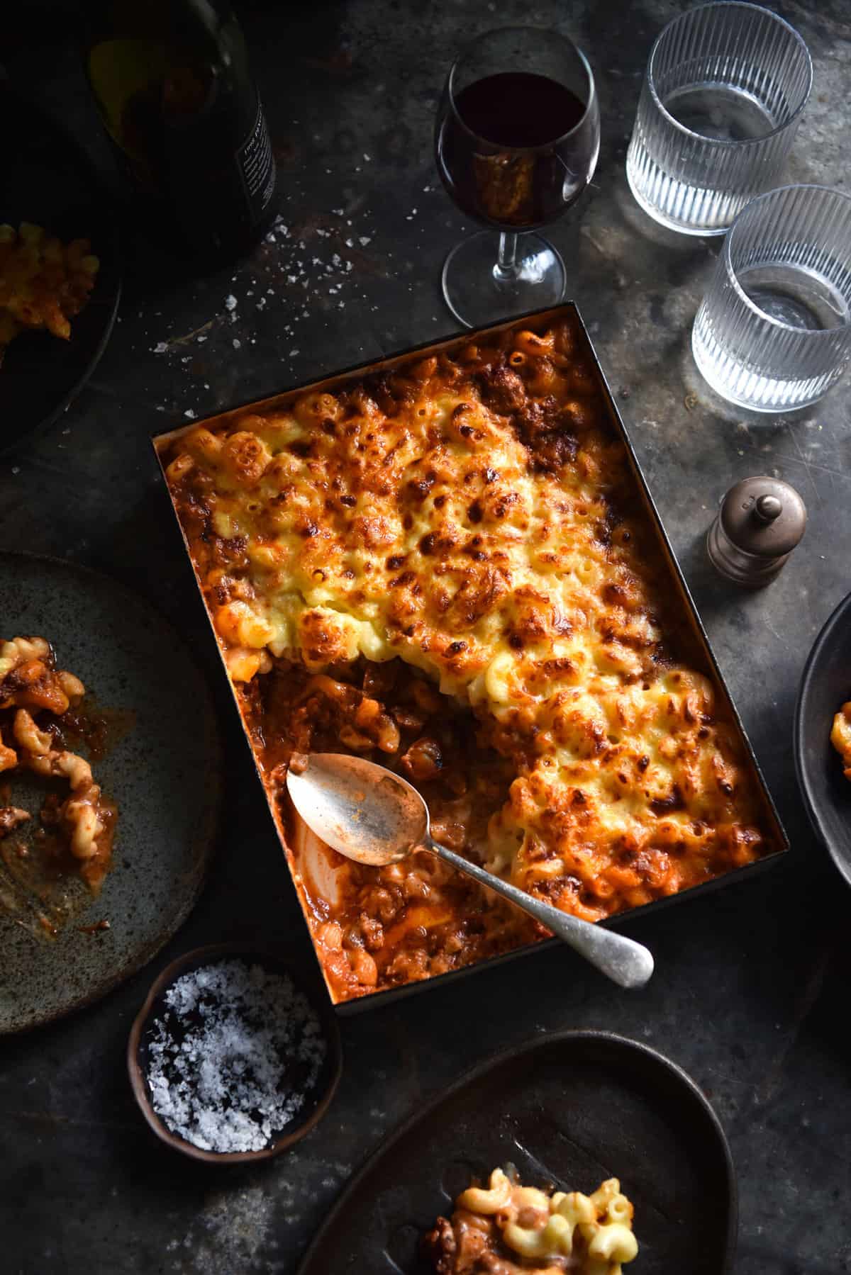 Gluten free, vegetarian bolognese, mac and cheese bake on a dark moody backdrop surrounded by water glasses. A scoop of bake has been removed, revealing the rich vegetarian bolognese underneath the mac and cheese