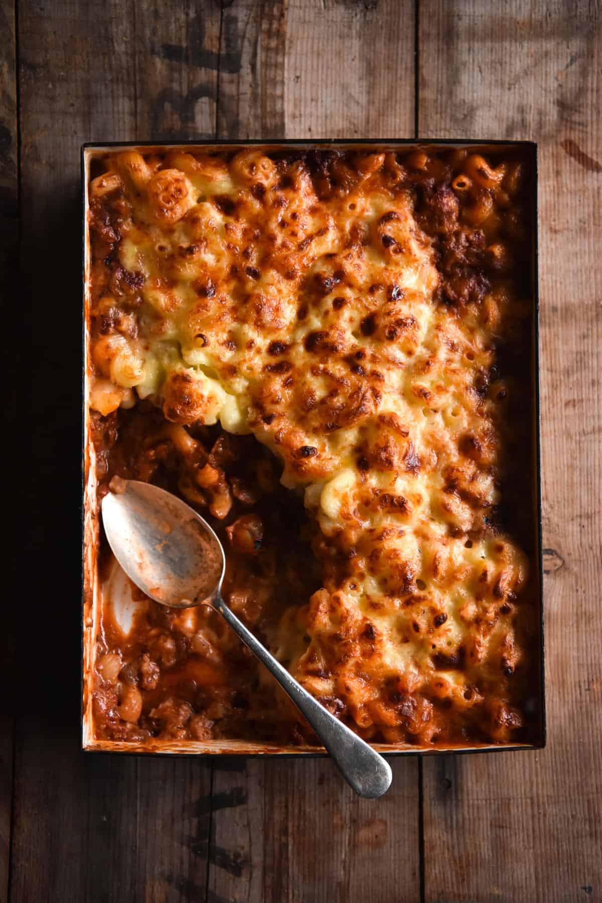 An aerial image of a vegetarian bolognese mac and cheese bake on a wooden table. A big spoonful of the bake has been removed and a vintage silver spoon sits in it's place.
