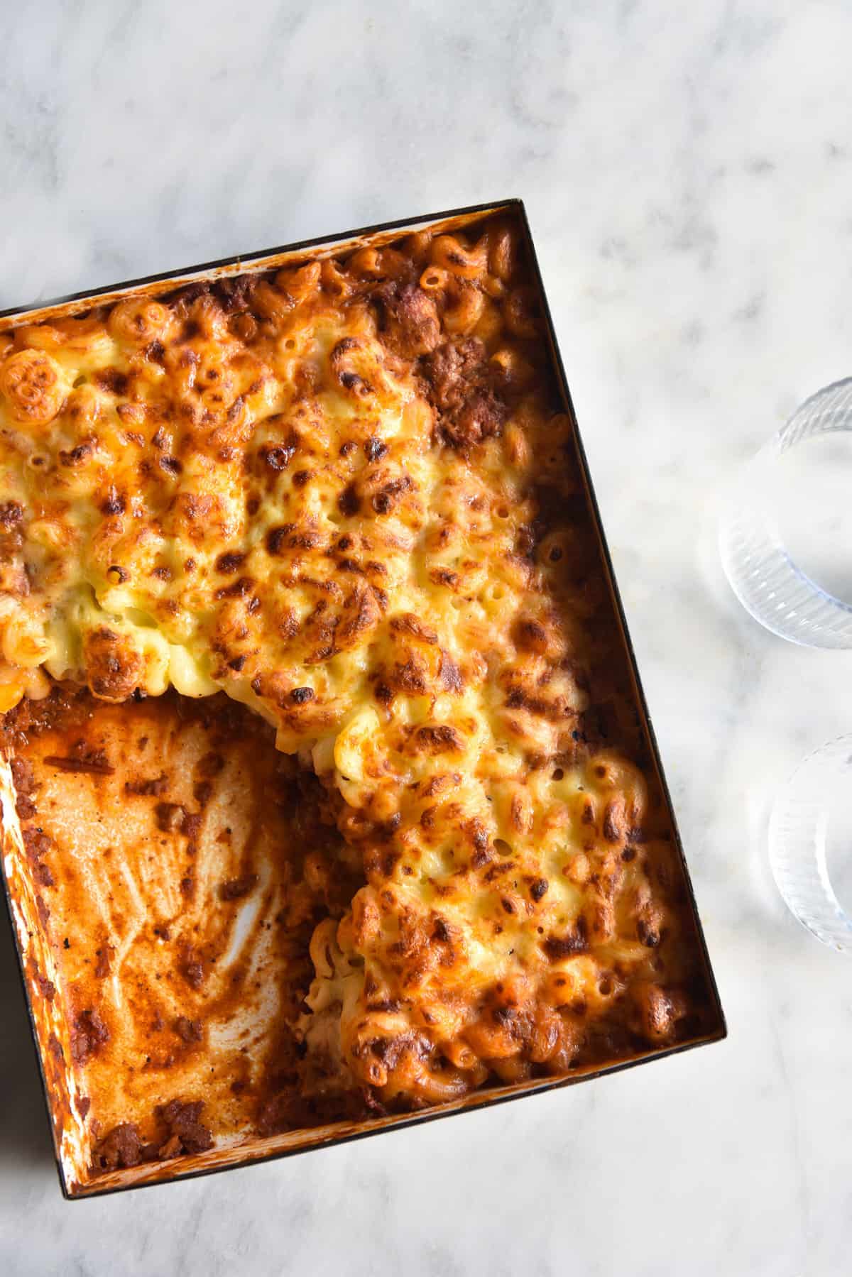 An aerial image of a vegetarian hamburger helper bake on a white marble table. A big scoop of the bake has been removed, revealing the rich mince underneath the creamy mac and cheese layer.