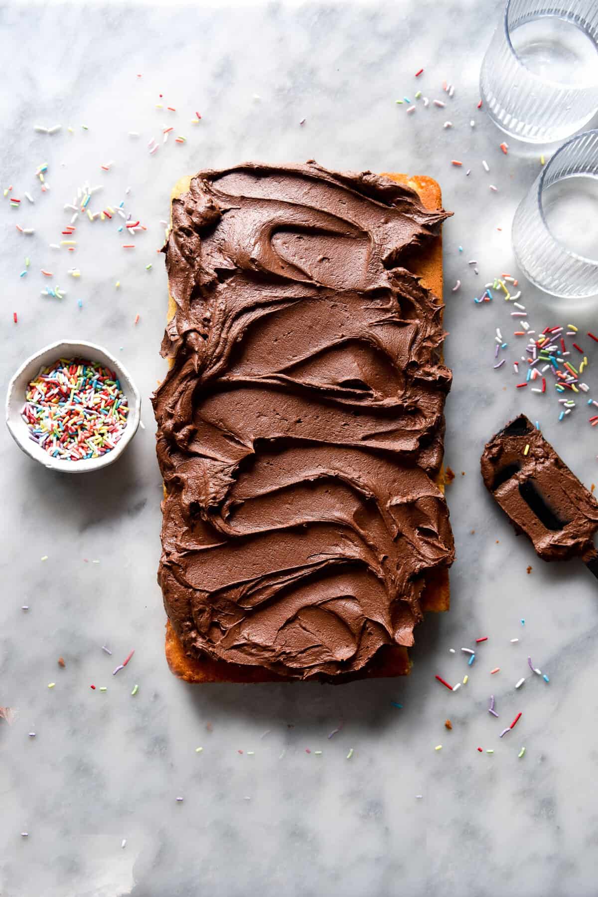 An aerial view of a brown butter chocolate buttercream topped cake surrounded by sprinkles on a white marble table. 