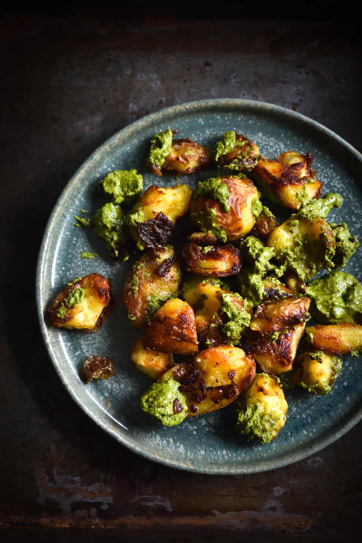 Crispy smashed potatoes with vegan rocket and almond pesto on a green ceramic plate against a dark steel backdrop