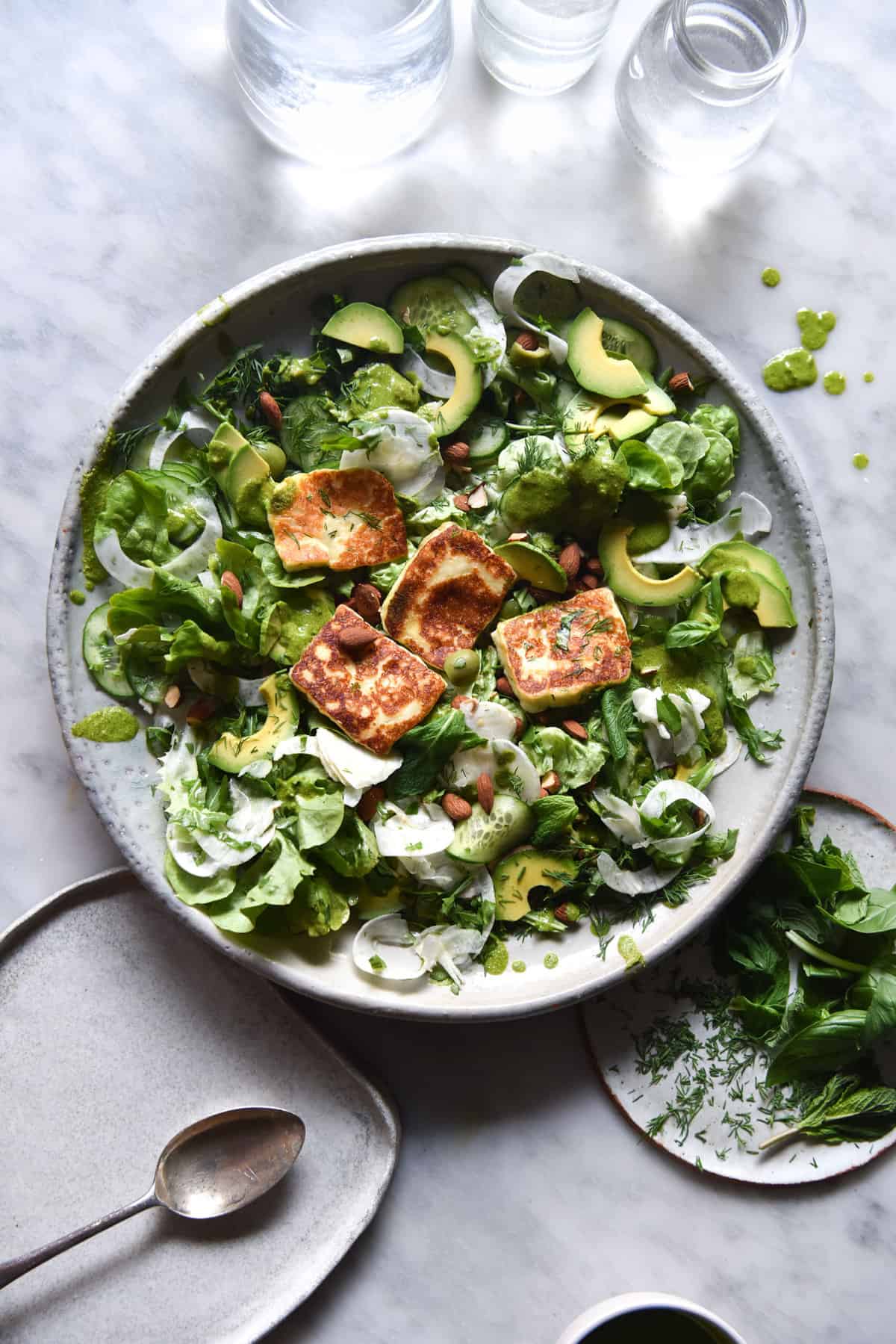 An aerial view of a halloumi, greens and pesto salad sitting atop a white marble table. Two glasses of water sit in the top of the image and create light and shadow patterns down onto the salad.