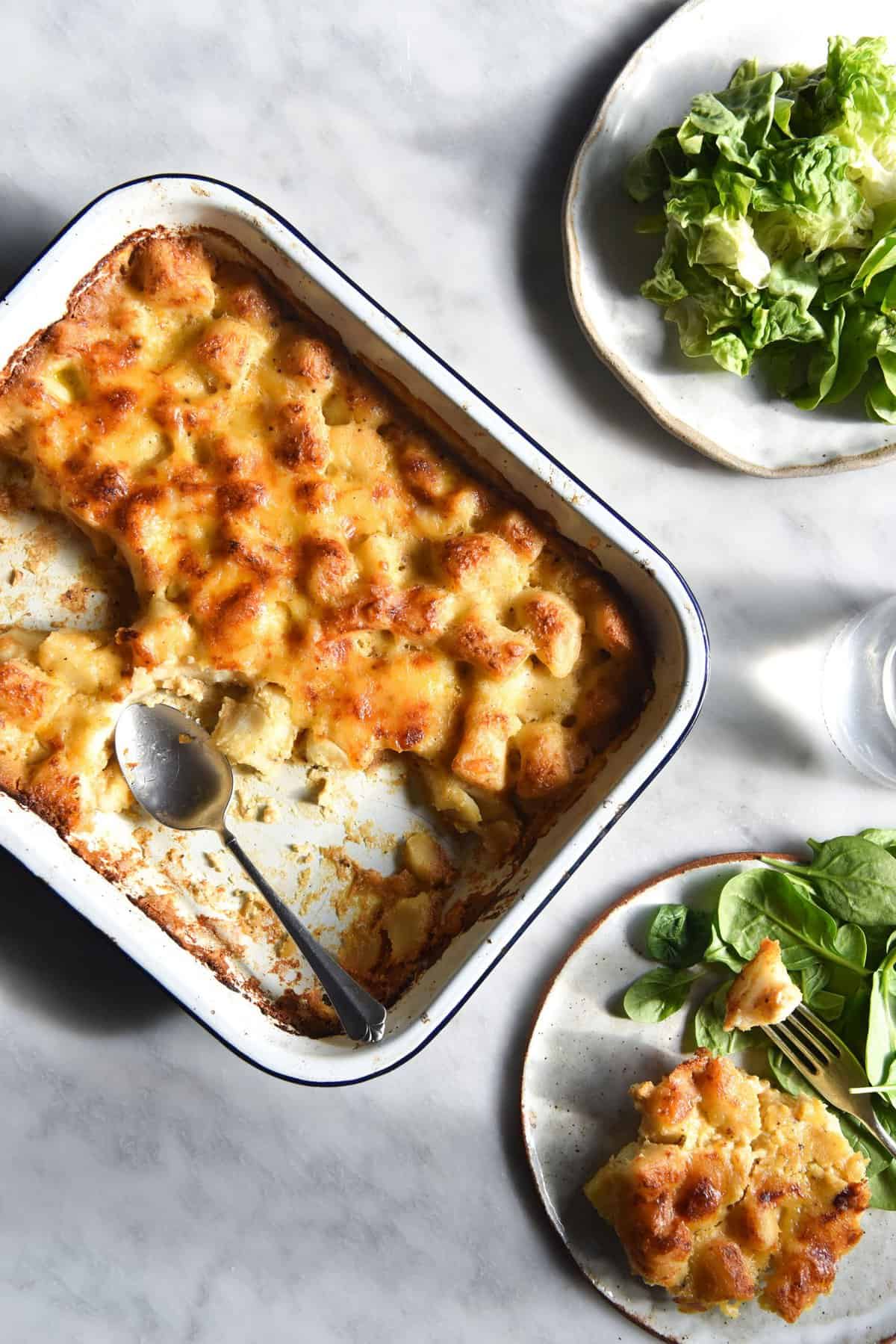 Gluten free gnocchi in a lactose free bechamel in a white oven dish. The dish sits on a white marble table and is surrounded by plates with gnocchi bake and salad on them.