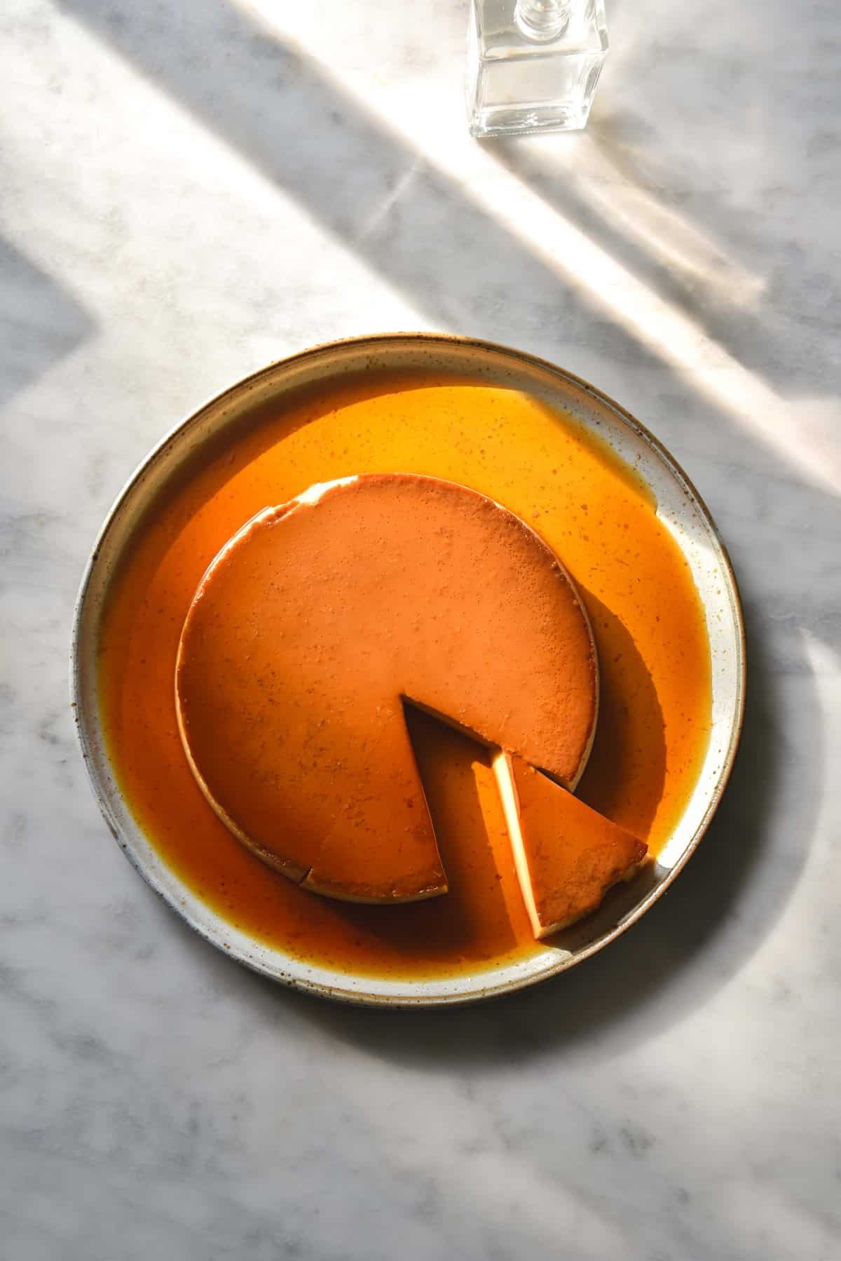 An aerial view of a sunlit white ceramic plate topped with a lactose free creme caramel. Light and shadow patterns from water glasses cast shapes across the top of the image