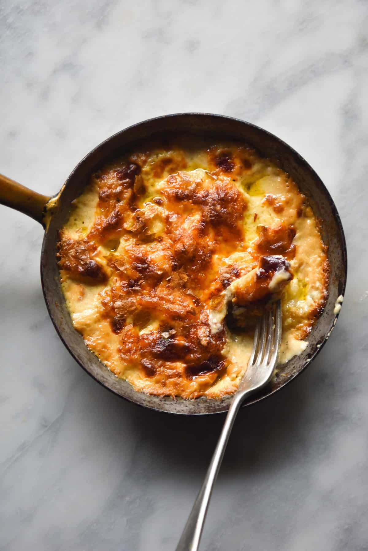 Gluten free gnocchi in a lactose free bechamel. The gnocchi has been baked in a small skillet and sits atop a white marble table. A fork sticks out of the bottom right corner of the image.