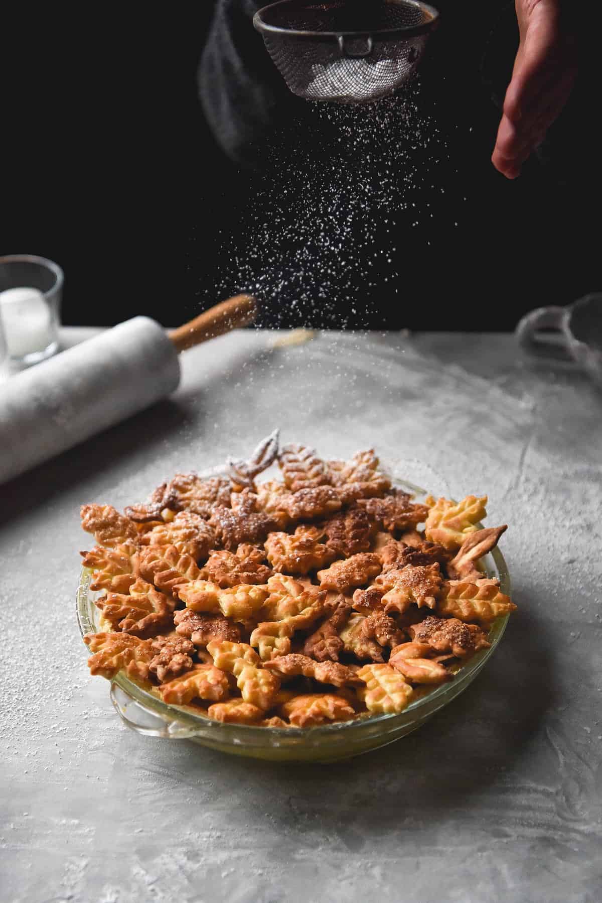 A gluten free pumpkin pie adorned with golden pastry leaves. In the background a hand is sprinkling icing sugar onto the pie.