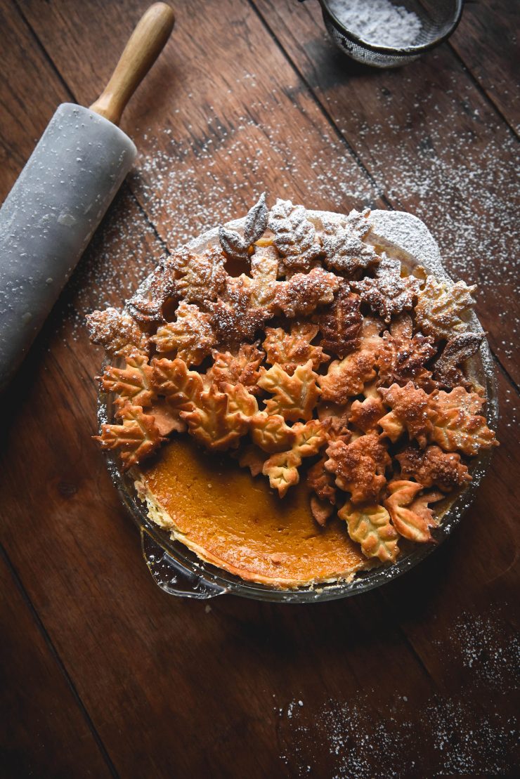 A gluten free pumpkin pie decorated with pastry leaves against a moody wooden backdrop