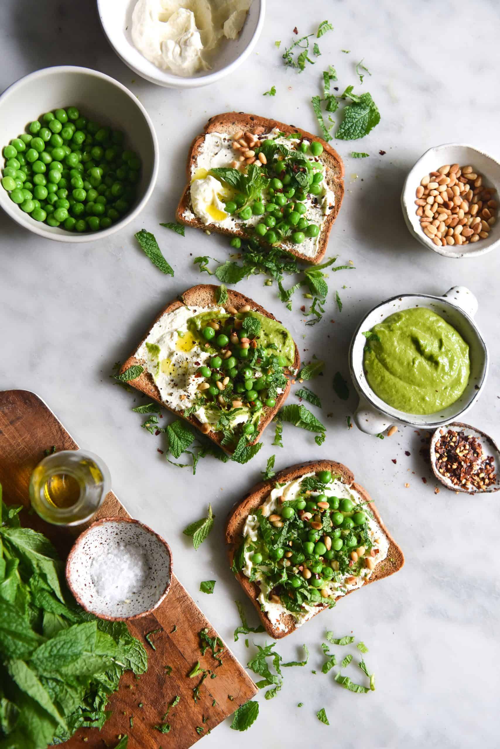 An aerial image of three pieces of gluten free toast casually strewn across a white marble table. The toasts are topped with lactose free ricotta, peas and mint. Surrounding the toasts are the ingredients used to assemble them, casually arranged. 