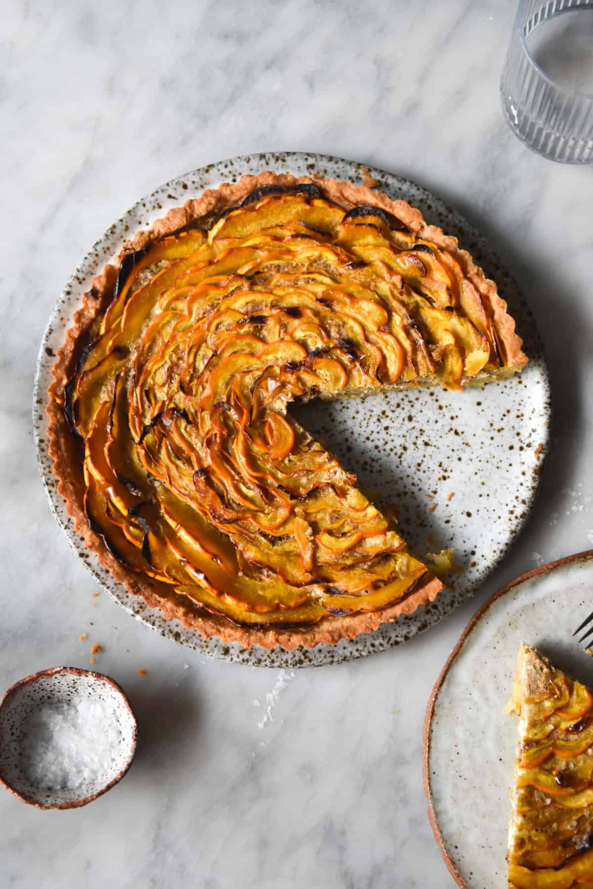 An aerial image of a savoury pumpkin tart on a white speckled ceramic plate atop a white marble table.
