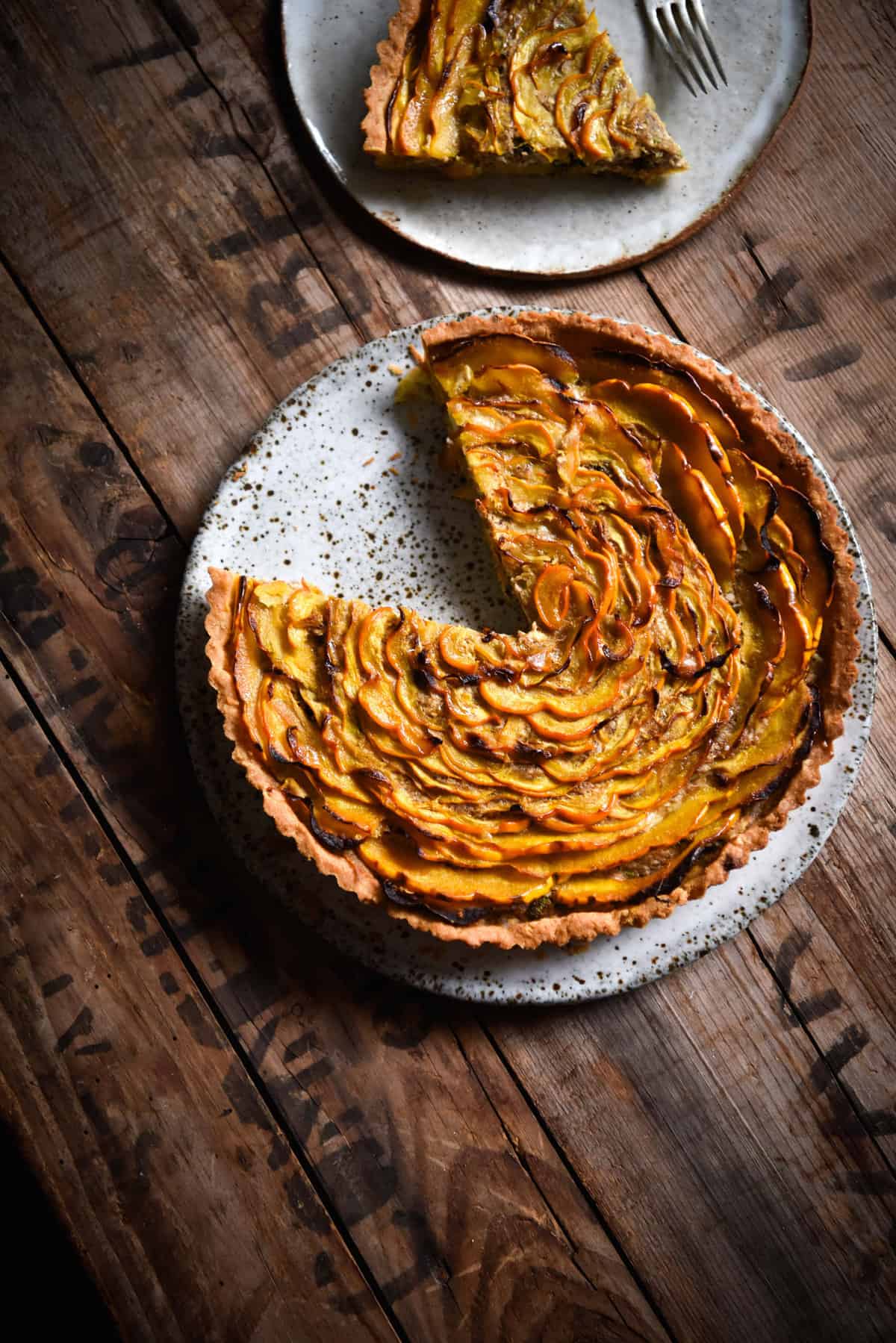 An aerial image of a gluten free savoury pumpkin tart on a white speckled ceramic plate atop a wooden table