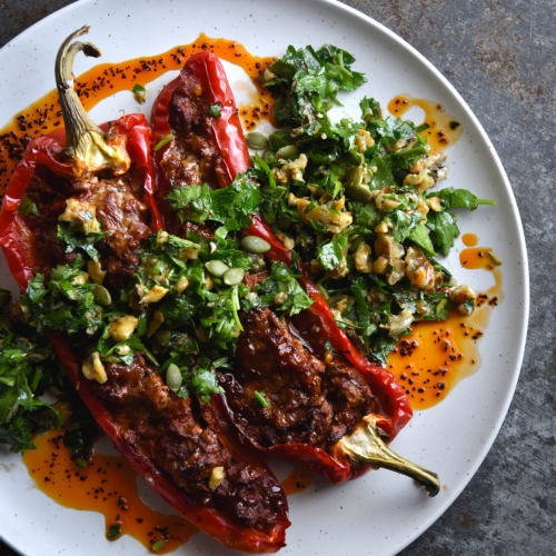 An aerial view of vegetarian mince stuffed bullhorn capsicums, topped with a walnut and parsley gremolata and doused in chilli oil. The vibrant feast sits atop a white plate, set against a mottled grey backdrop