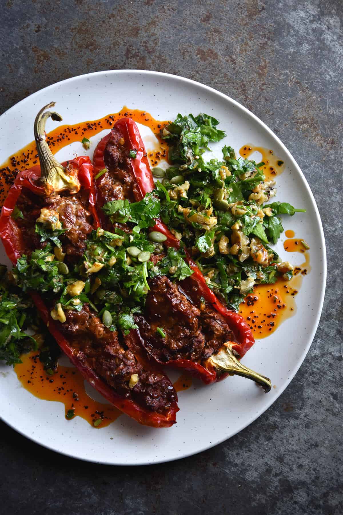 An aerial view of vegetarian mince stuffed bullhorn capsicums, topped with a walnut and parsley gremolata and doused in chilli oil. The vibrant feast sits atop a white plate, set against a mottled grey backdrop