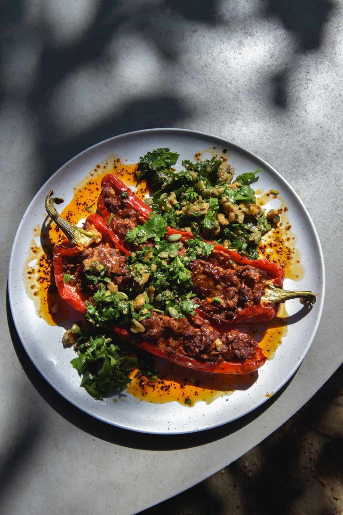 An aerial view of FODMAP friendly vegetarian mince stuffed bullhorn capsicums topped with herb and walnut gremolata and doused in chilli oil. The capsicums sit atop a white plate on an grey outdoor table which sits in contrasting, dappled sunlight