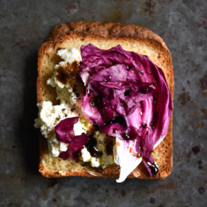 An aerial image of a piece of toast on a dark grey mottled backdrop. The toast is topped with lactose free ricotta, radicchio and a caramelised balsamic glaze.