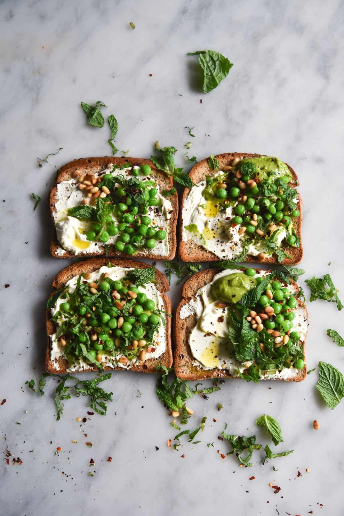 An aerial image of four pieces of toast atop a white marble table, arranged into a square. The toasts are topped with lactose free ricotta, peas, mint and chilli flakes.