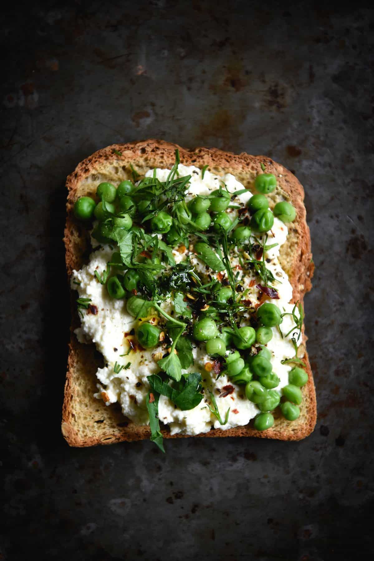 An aerial image of a piece of toast on a mottled grey backdrop. The toast is topped with smashed peas, herbs, chili flakes and lactose free ricotta.