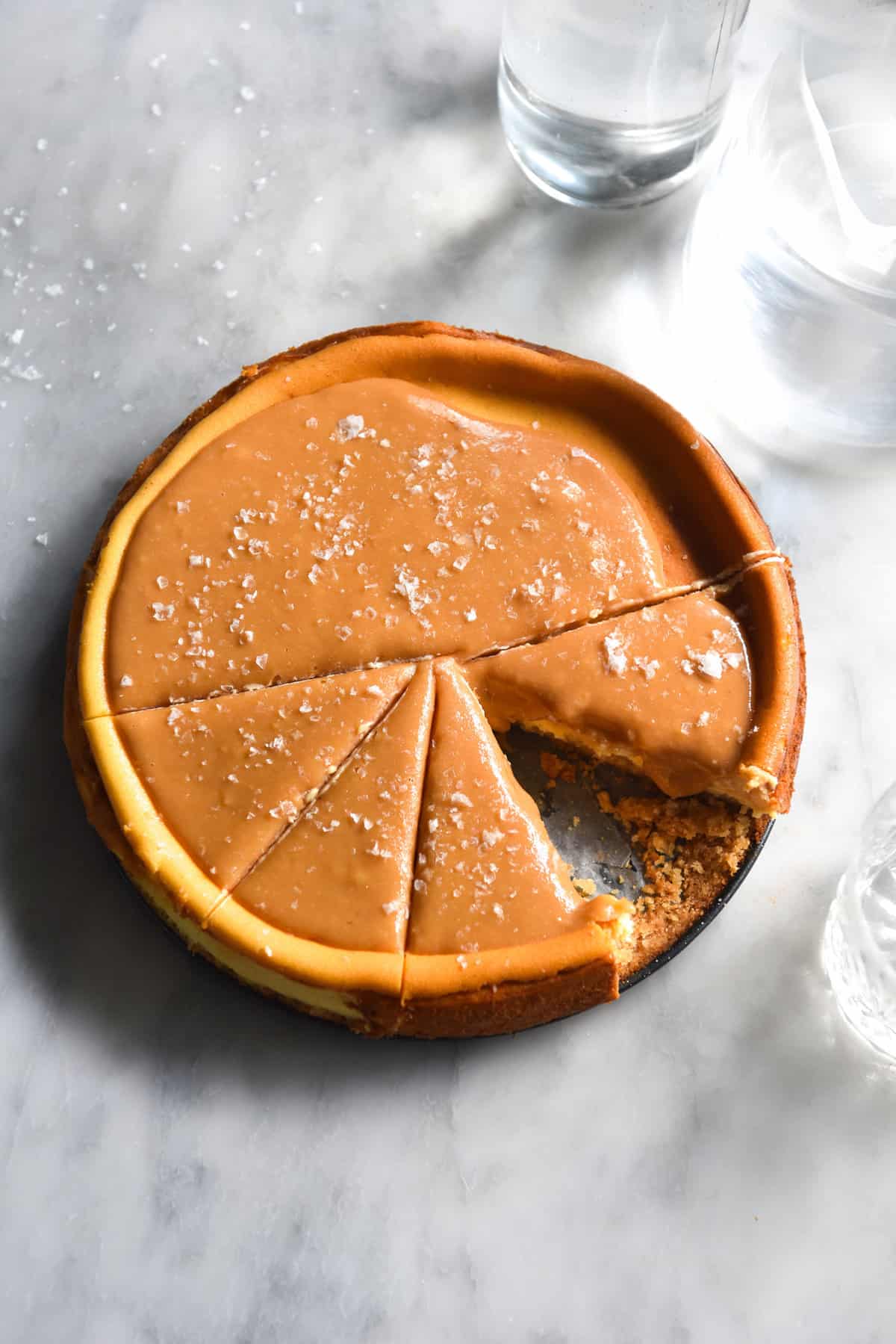 An aerial image of a lactose free cheesecake on a gluten + grain free base with lactose free salted caramel atop a white marble table. The caramel top of the cheesecake is finished with sea salt flakes. 