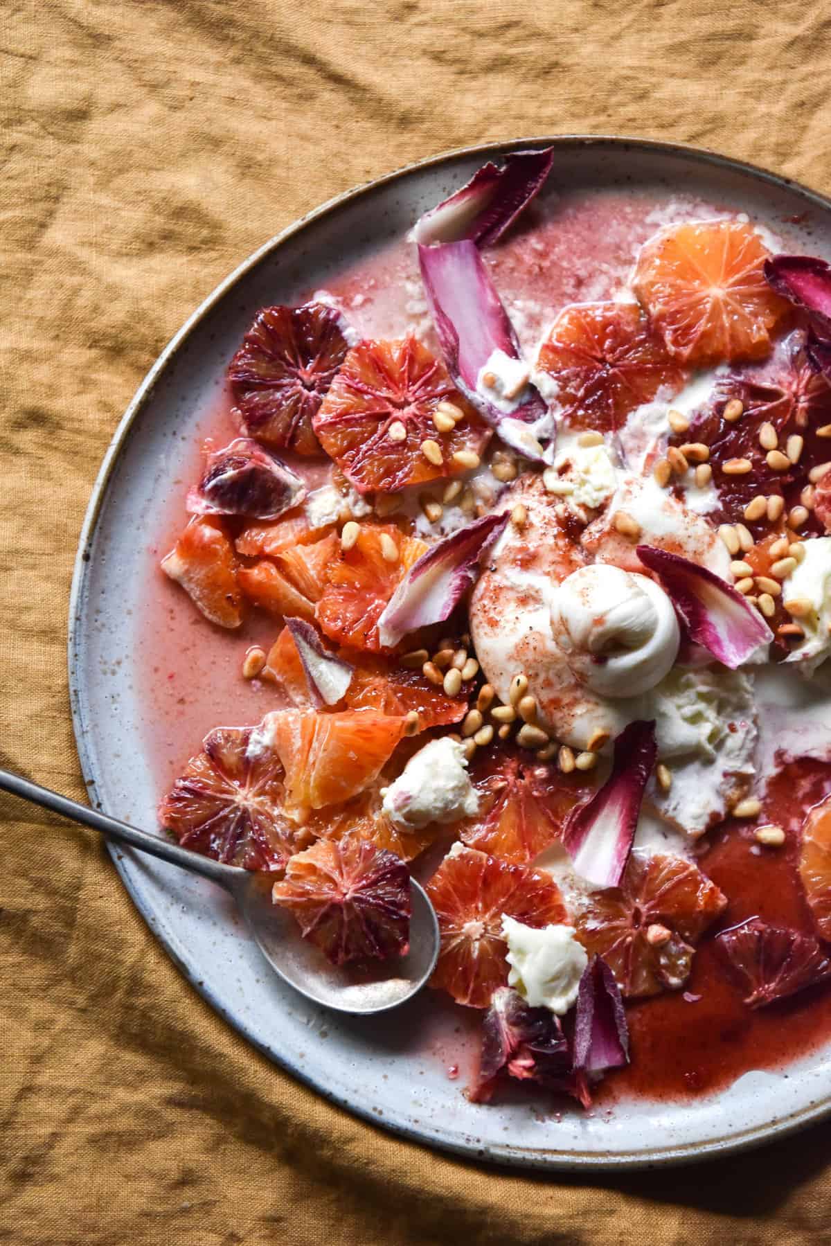 An aerial image of a white ceramic plate topped with a burrata, orange, radicchio and pine nut salad atop a turmeric coloured linen tablecloth.