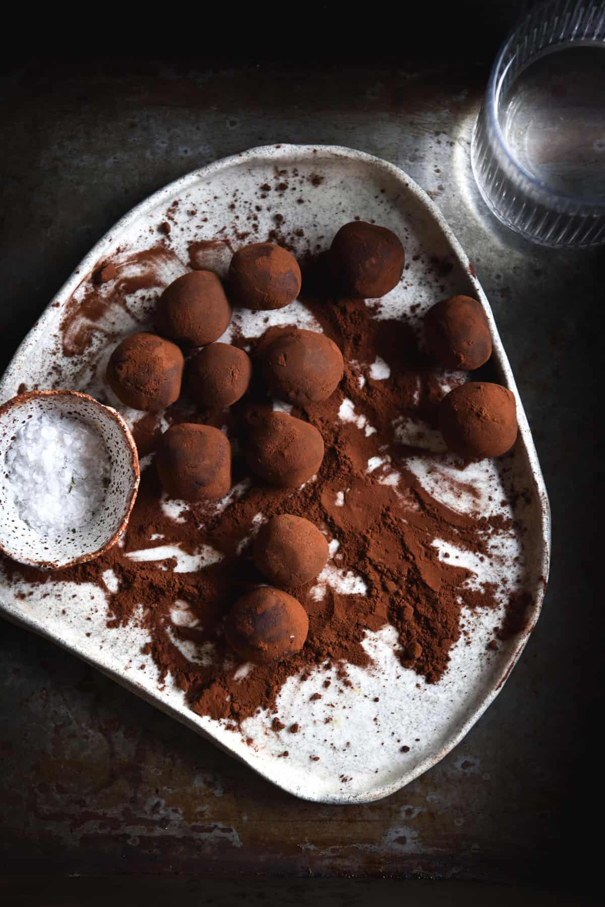 An aerial moody image of vegan Nutella truffles on a white ceramic plate against a dark steel backdrop. The truffles are covered in cocoa and a pinch bowl of sea salt sits to the left of them,