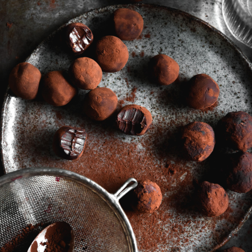 Vegan 'nutella' truffles on a dark moody backdrop and a dark grey ceramic plate. The truffles have been dusted with cocoa, and a sieve with cocoa sits to the bottom left of the image