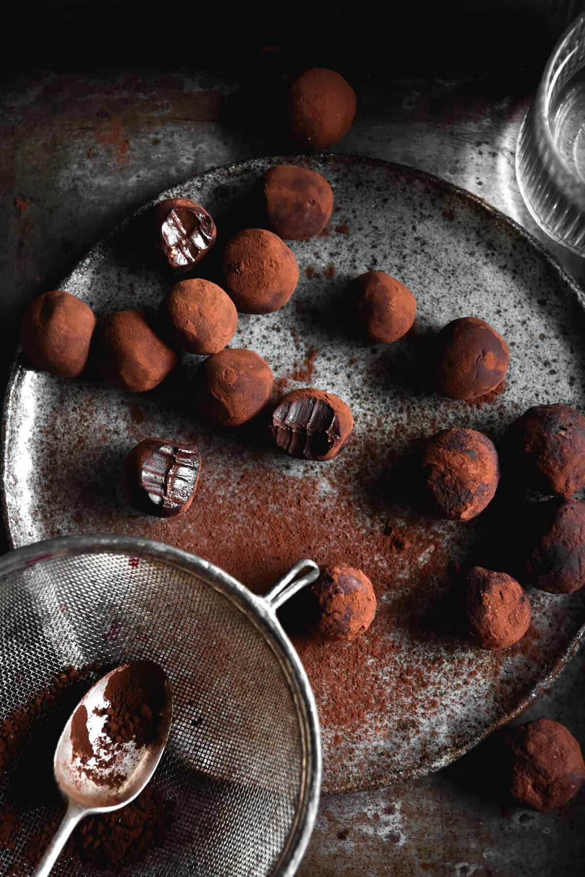 Vegan 'nutella' truffles on a dark moody backdrop and a dark grey ceramic plate. The truffles have been dusted with cocoa, and a sieve with cocoa sits to the bottom left of the image
