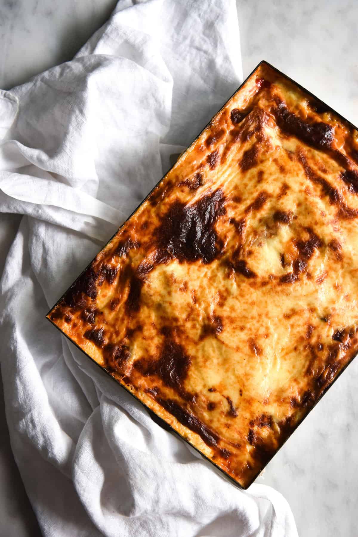 An aerial view of a golden brown vegan gluten free lasagne atop a white marble table and a white linen cloth.