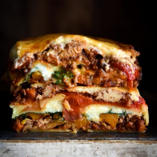 A side on image of a gluten free lasagne stack against a black backdrop. The lasagne is layered with vegetarian ragu, roasted vegetables, paste sheets and a gooey bechamel.