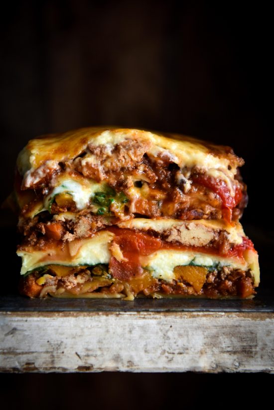 A side on image of a gluten free lasagne stack against a black backdrop. The lasagne is layered with vegetarian ragu, roasted vegetables, paste sheets and a gooey bechamel.