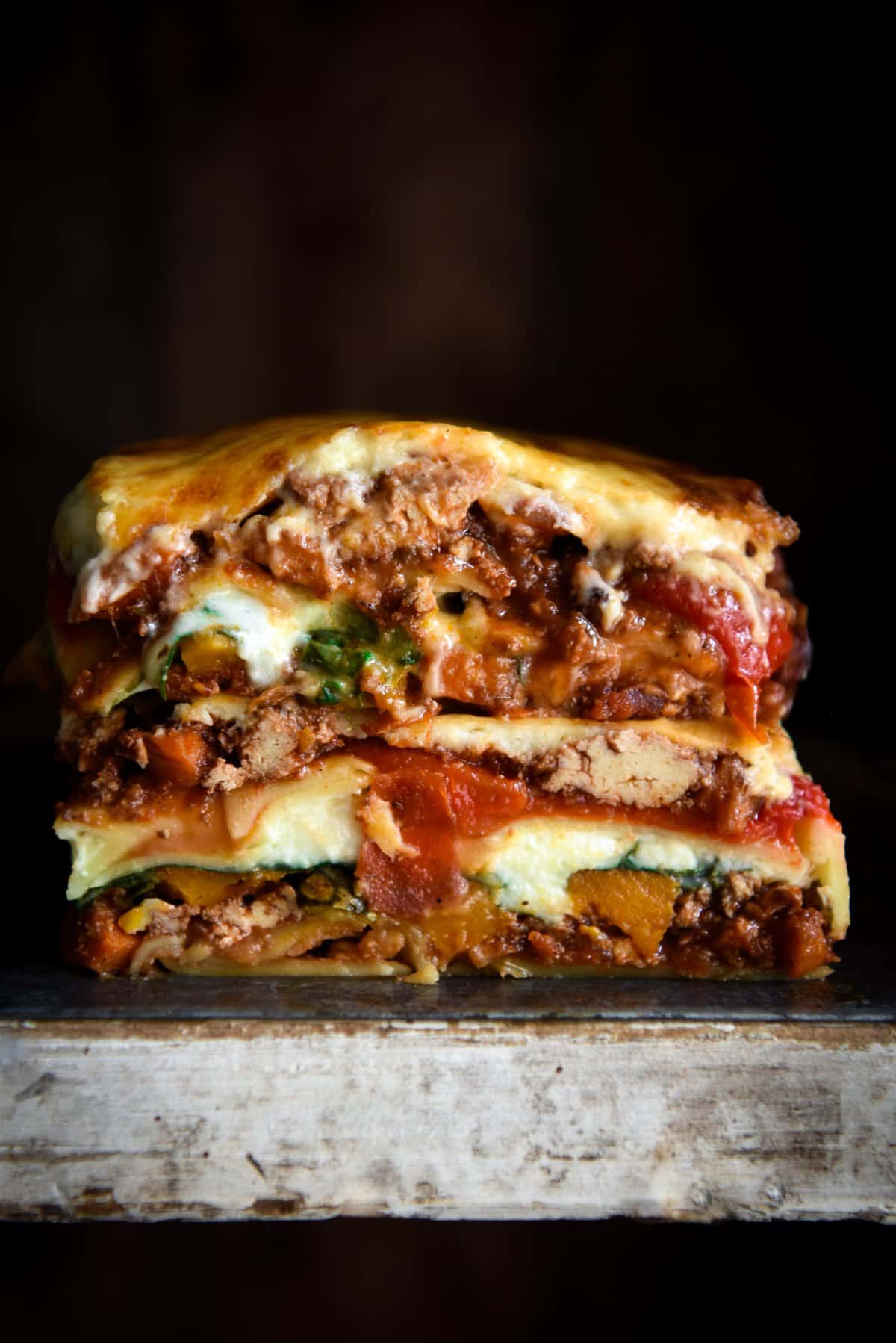 A side on image of a gluten free lasagne stack against a black backdrop. The lasagne is layered with vegetarian ragu, roasted vegetables, paste sheets and a gooey bechamel. 