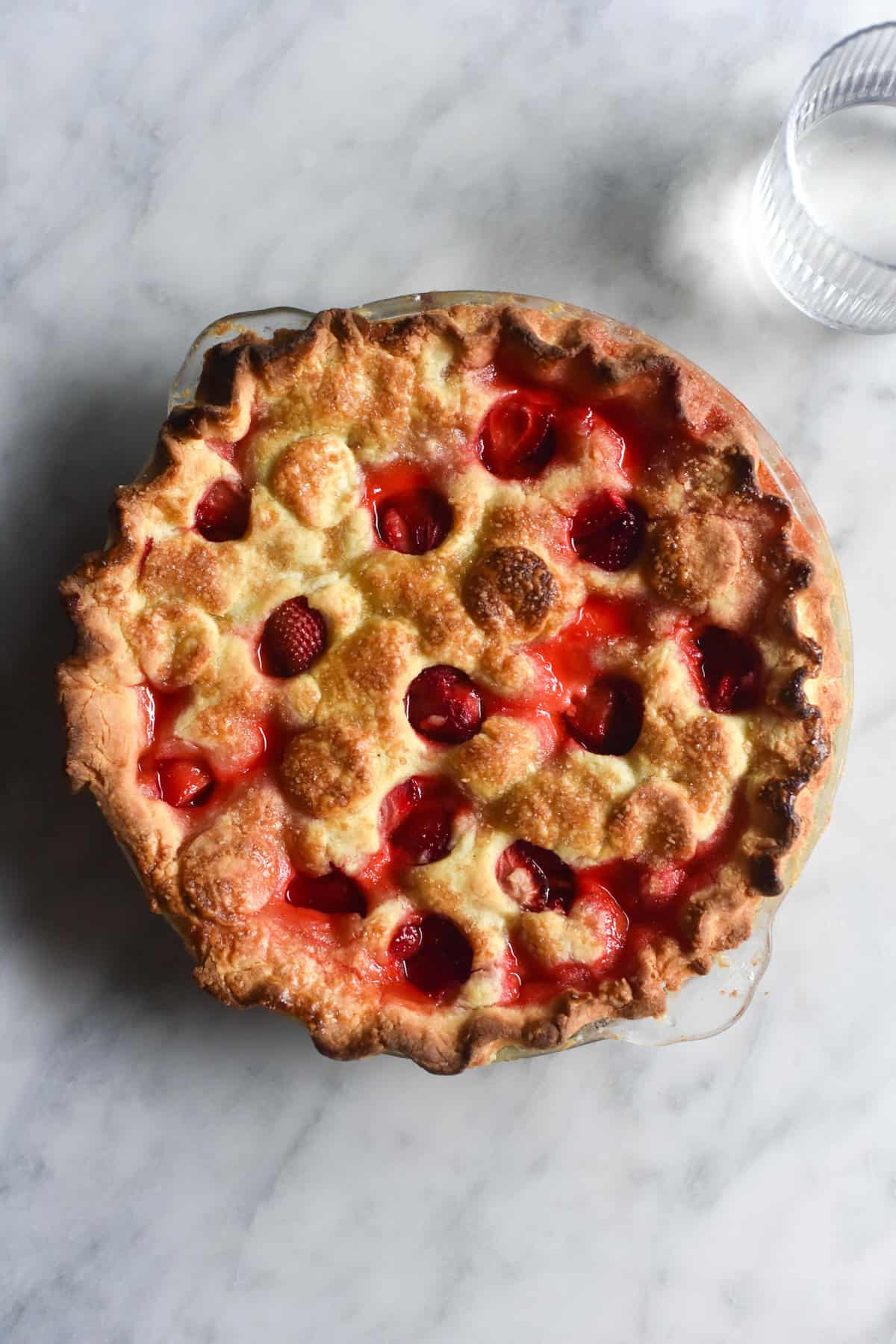 Gluten free strawberry pie on a white marble table. The pastry is golden brown and has holes of pastry removed in a pattern, revealing the cooked strawberries underneath. 