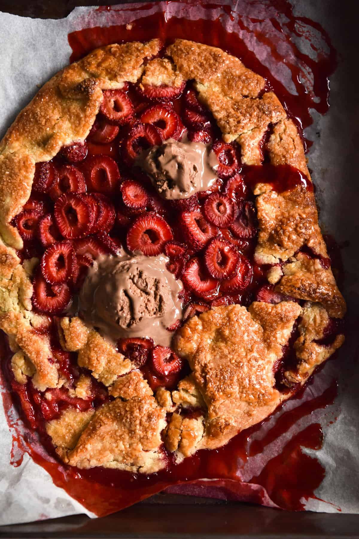 Gluten free strawberry galette on a piece of baking paper. The galette is golden brown and leaking strawberry juices. It is topped with melting chocolate ice cream.