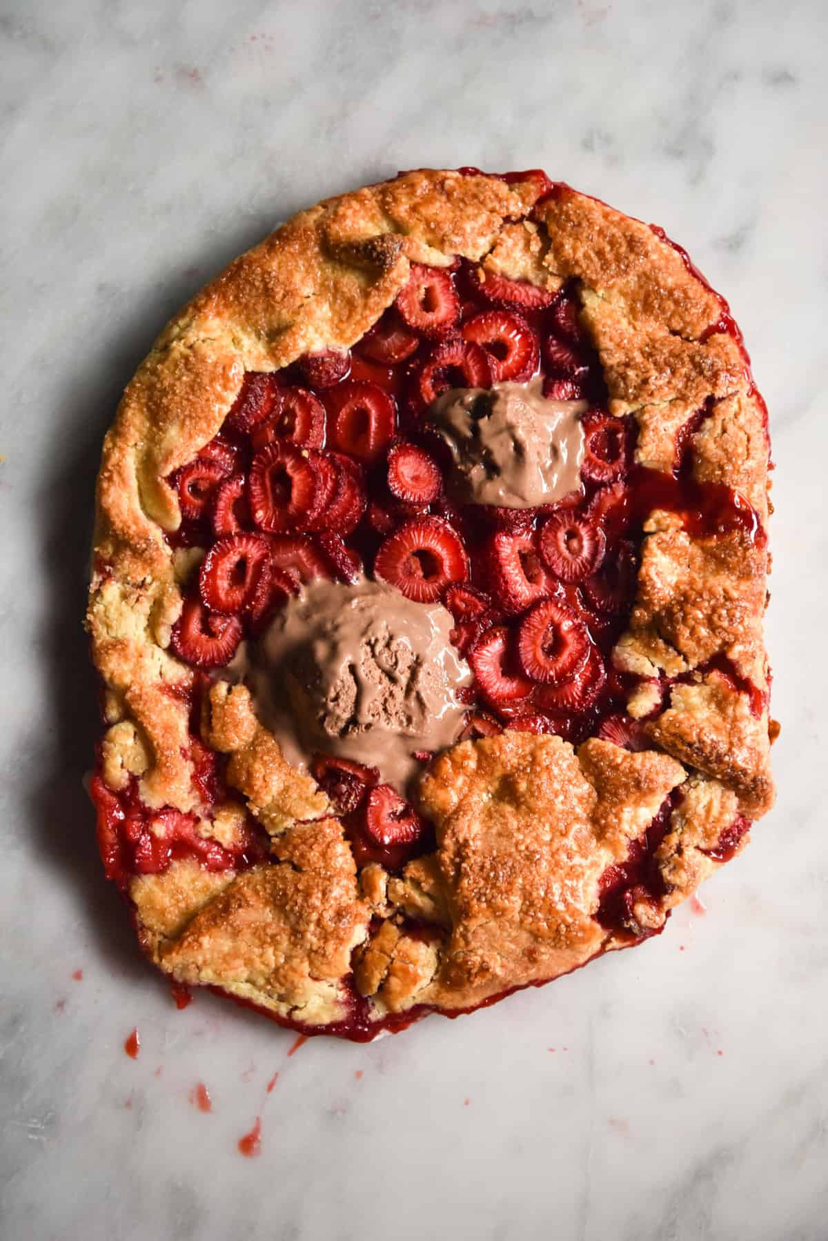 An aerial view of a gluten free strawberry galette on a white marble table. The galette is topped with melty chocolate ice cream. 