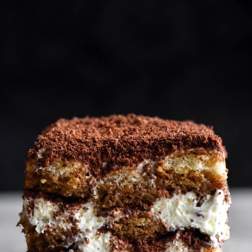 A close up, side on view of a slice of gluten free, FODMAP friendly tiramisu. The slice sits atop a marble table which contrasts with a black background