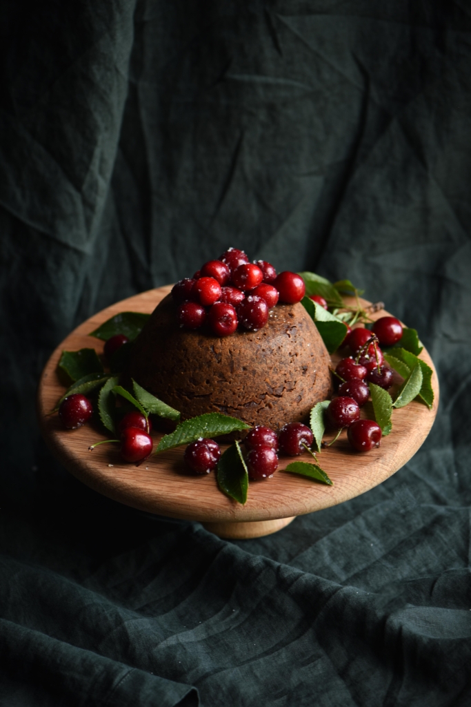 A fruitless, gluten free christmas pudding covered in cherries against a dark green backdrop