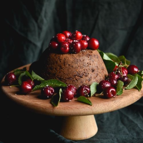 A side on view of a fruitless Christmas pudding topped with cherries against a green backdrop